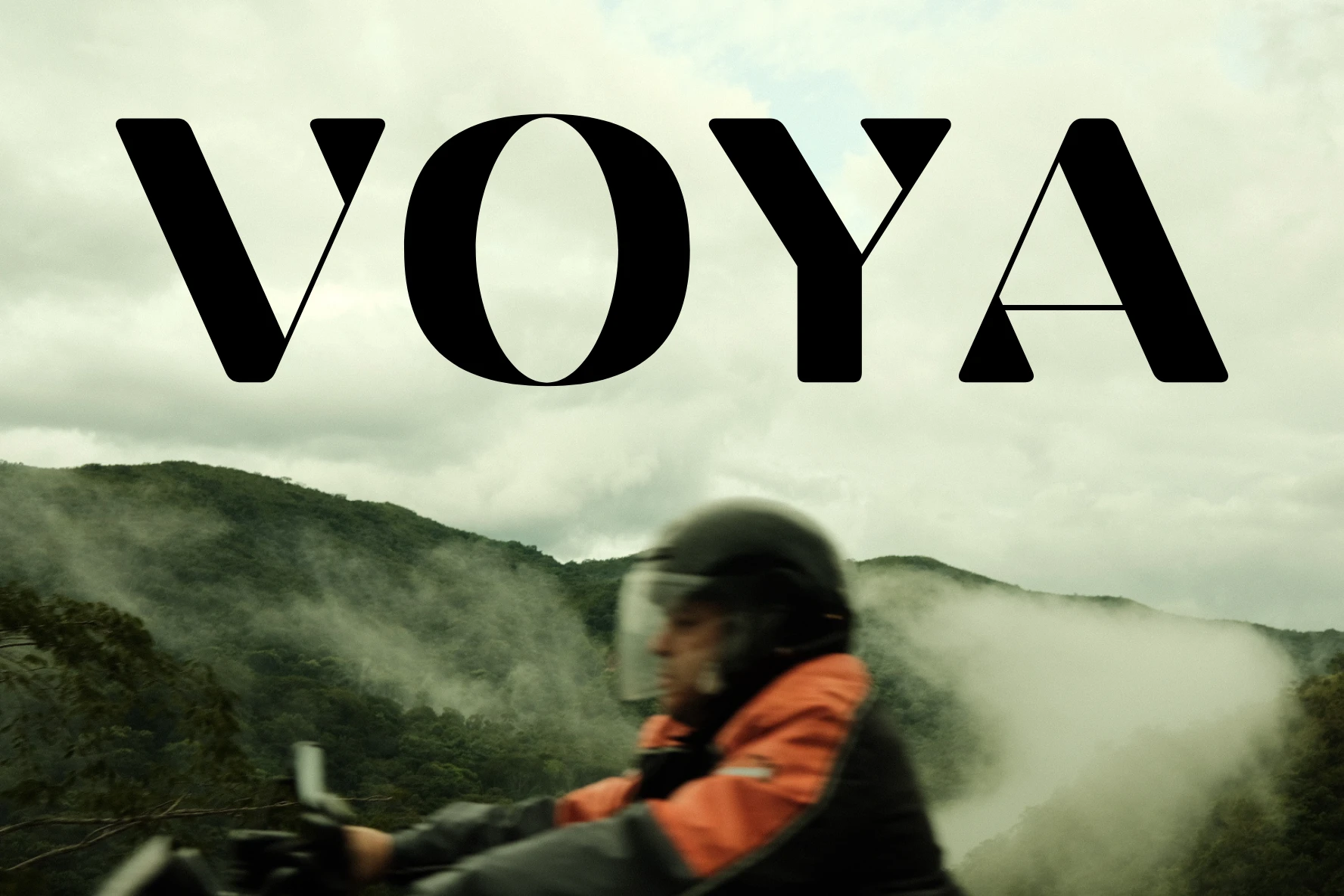 Motociclista con chaqueta naranja cruza una montaña cubierta de neblina, bajo el logotipo VOYA
